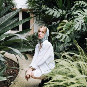 A young woman in a white blouse with a headscarf poses in a lush garden setting.