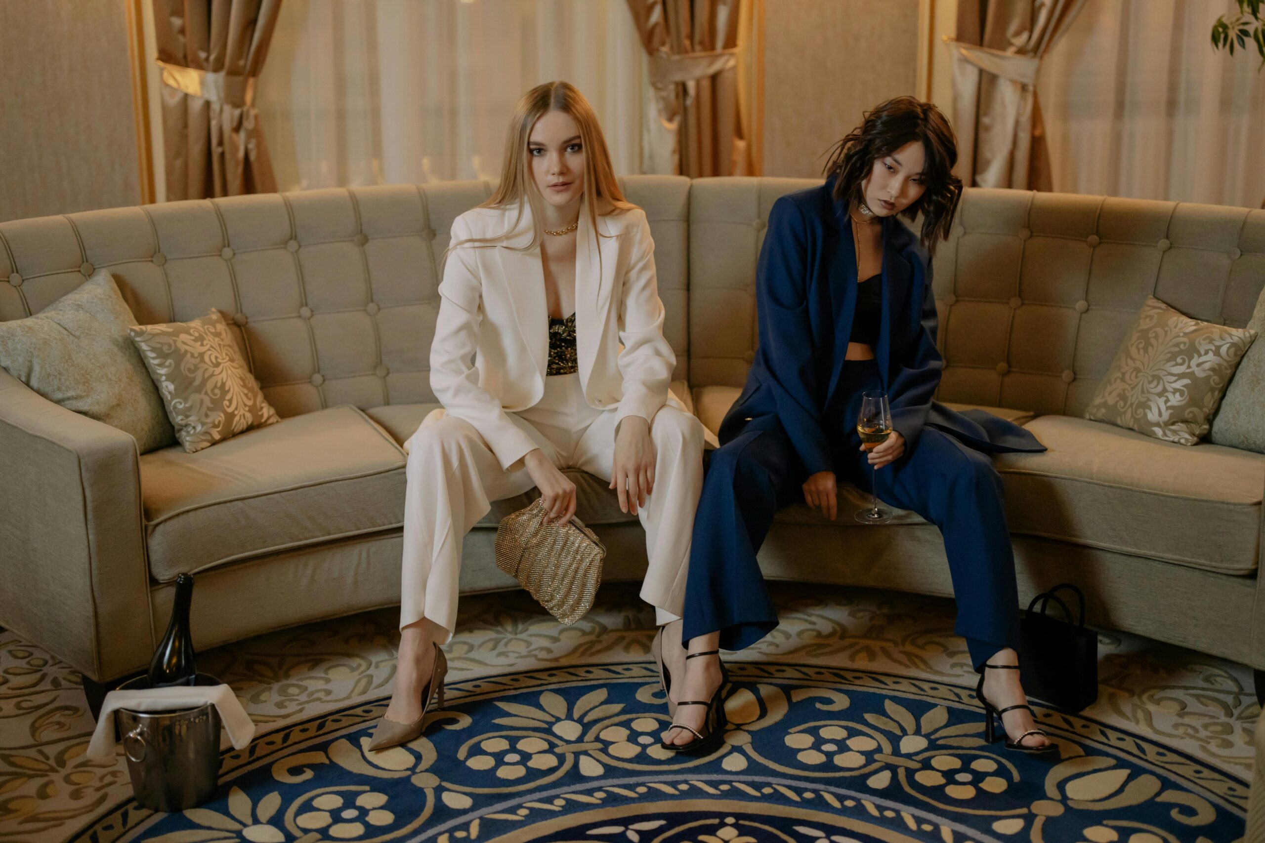 Two women in luxurious suits sit on a sofa holding champagne, exuding elegance and glamour.