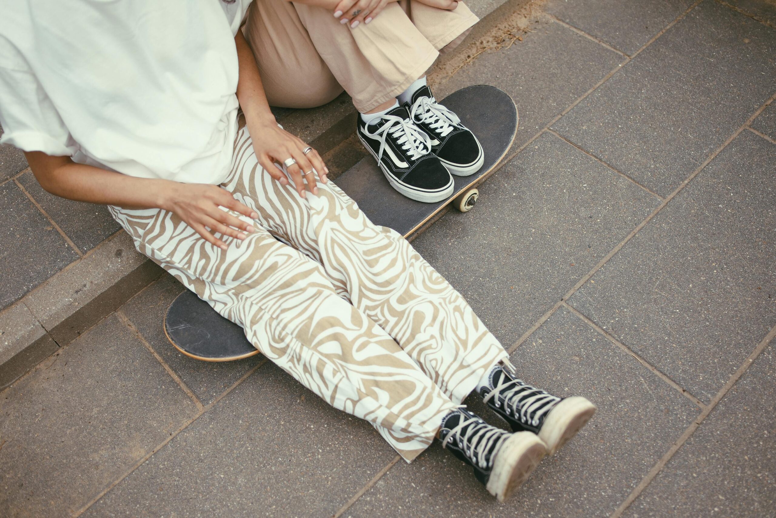 Two people in fashionable pants and sneakers sitting on skateboards outdoors on a paved surface.